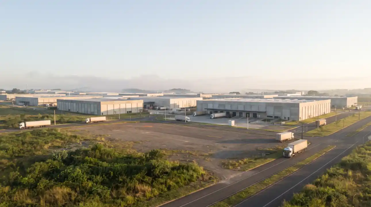Vista aérea de um complexo industrial com galpões, pátios e vias de acesso, ao amanhecer, com caminhão estacionado em uma estrada interna.